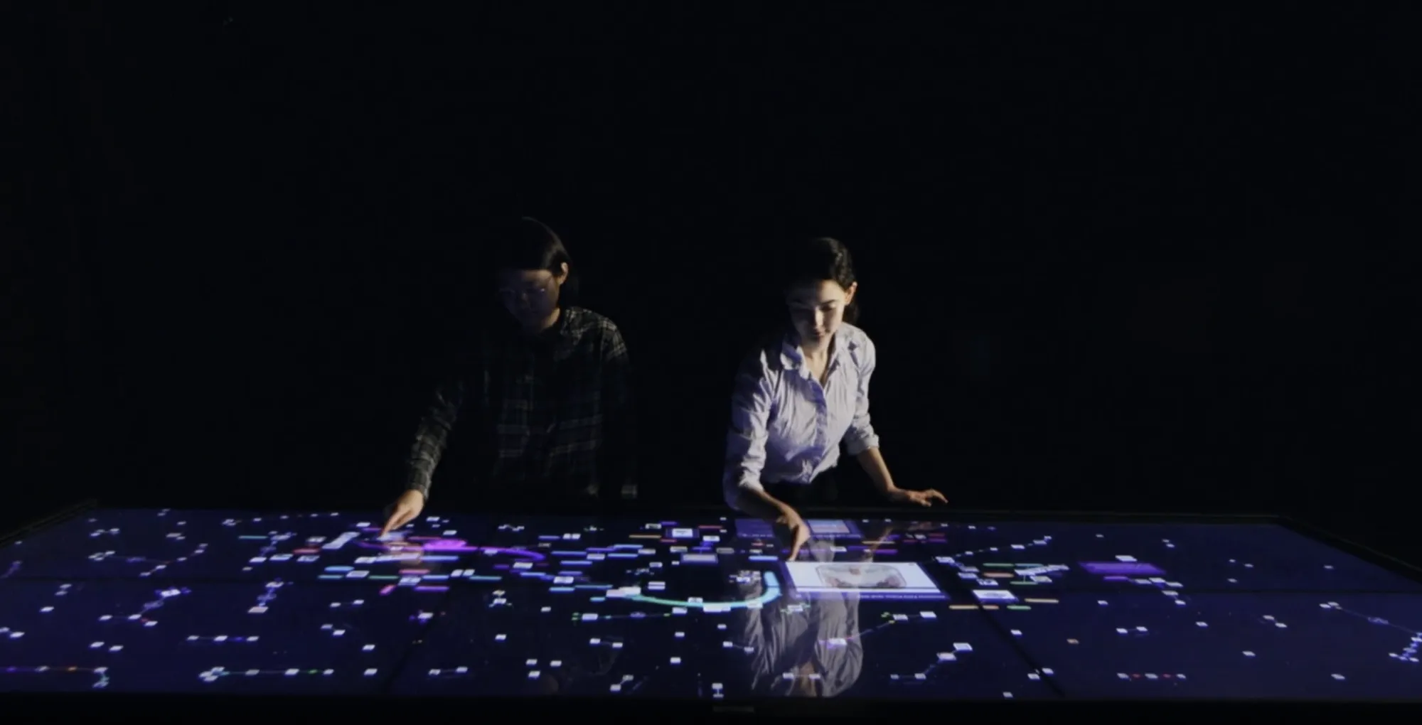 Two visitors exploring glowing museum objects on the AI Connections Table in a dark gallery.